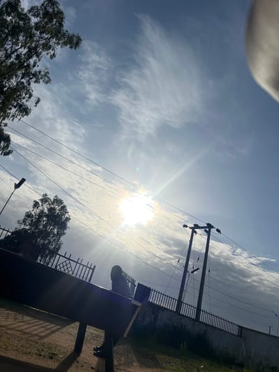 The image shows a bright, sunny day in Ban, Nigeria, with the sun shining strongly through a partly cloudy sky. The sun's rays create a lens flare effect. In the foreground, a person is partially visible, appearing to be standing near a dark, rectangular structure with legs, possibly a bench or table. The person is silhouetted against the bright sky and is wearing dark clothing. A dark fence with a patterned grill runs along the right side of the frame, separating the area from a patch of grass and dirt. Power lines and a utility pole are prominent in the sky, stretching across the frame. In the background, trees with green foliage are visible on the left and right sides of the image. The overall scene suggests an outdoor environment, possibly a park or a residential area, during daylight hours. The weather appears to be warm and clear, despite the presence of some clouds. No visible text is present in the image.