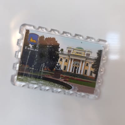 A clear plastic souvenir magnet, rectangular with a serrated border resembling a postage stamp, is depicted lying on a white, reflective surface. The magnet features a color photograph of a prominent, multi-story palace characterized by a yellow facade, white classical columns supporting a central portico, and a distinct green domed roof. In the foreground, a circular fountain actively sprays water, positioned near a dark, seated figure statue atop a rectangular pedestal. The immediate surroundings consist of manicured green lawns interspersed with beds of red and orange flowers. Dense green trees frame the palace on both sides. In the upper left quadrant of the photograph, a blue heraldic shield with a golden reclining lion is visible, beneath which the white Cyrillic text "Гомель" (Gomel) is printed. Reflections of ambient light are visible on the magnet's plastic surface and the underlying white background. The scene depicted on the magnet is of the Rumyantsev-Paskevich Palace in Gomel, Belarus.