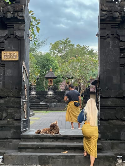 In Dawan, Indonesia, a group of people and a dog are at the entrance of a stone temple complex. A man wearing a dark t-shirt and a yellow sarong, and a woman with long blonde hair also wearing a yellow sarong and a blue shirt, are walking through a large stone gateway. A brown, shaggy dog is lying on the steps in front of the gateway, looking up at the people. The temple complex features dark stone architecture, ornate gateways, and traditional Balinese structures. The sky is overcast, and lush green trees surround the area. A sign with "NGERANJING MASUK" is visible on the left side of the gateway.