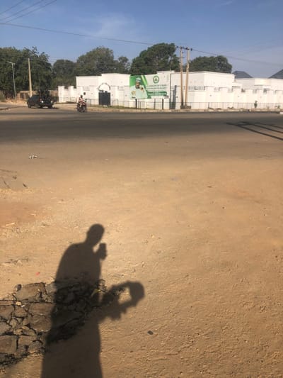 A ground-level, slightly off-kilter shot depicts a dusty road with a blurry shadow of a person holding a camera in the foreground. Adjacent to the road, white buildings line the left side, punctuated by trees and a billboard. A few motorcycles are visible in the distance. The sky is light blue and mostly clear.