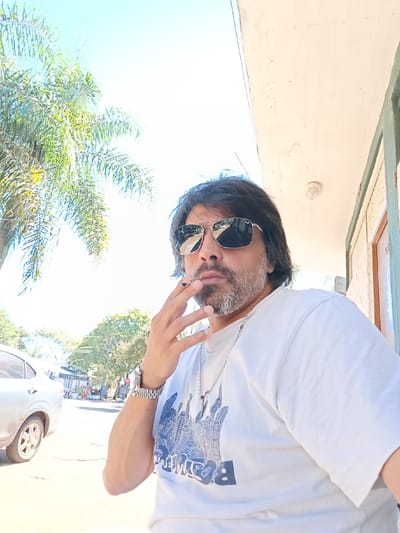 A man wearing sunglasses and a white t-shirt is smoking, with a tree and a building visible in the background. The image was taken in the city of Termas del Daymán, Uruguay. A silver car is visible on the left. The man is wearing a silver watch and a silver necklace with a pendant. He has dark hair and a salt-and-pepper beard. The weather appears to be sunny.