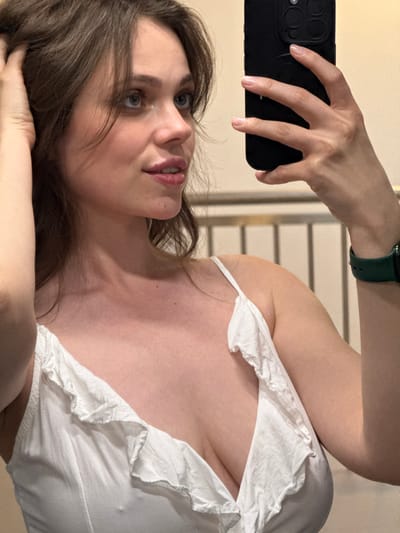 A fair-skinned woman with brown hair is holding a black smartphone up, presumably taking a selfie. She is wearing a white top with ruffles around the neckline. A green smartwatch is visible on her left wrist. The background includes a partially visible wall or room with horizontal lines. The location is not discernible as Balatonlelle, Hungary from the provided image.