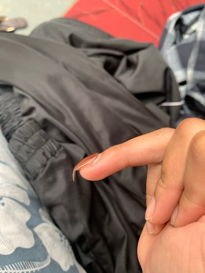 A close-up shot focuses on a person's index finger with a long, tan-colored artificial nail. The finger is extended towards the viewer, and the nail appears to be slightly curved at the tip. In the background, a dark-colored jacket is draped, with visible wrinkles and a cinched waistband. To the right of the jacket, a portion of a plaid shirt is visible. The lower left of the image shows a blue and white patterned fabric, possibly a blanket or pillow. The scene is indoors, with soft lighting.