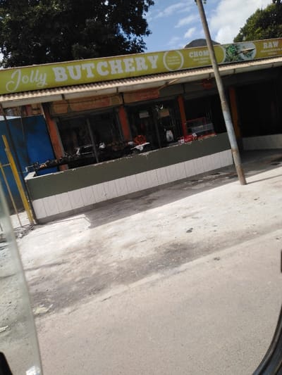 The image displays the exterior of "Jolly BUTCHERY" located in Kikuyu Township ward, Kenya. The establishment features a prominent yellow signboard with the business name in white text, accompanied by additional text reading "The Meat that serves happiness," "CHOM (Beef, mutton & Chicken)," and "RAW (Minced Mutton & Chicken)." The shopfront consists of multiple large glass windows, displaying an "OPEN" sign and two contact telephone numbers: 0721 977 749 and 0704 63 29 11. Below the windows, a low wall is visible, constructed with dark green tiles on the upper section and white tiles on the lower section. The foreground is occupied by a grey, unpaved road surface. A tall, slender utility pole stands vertically in the mid-ground to the right, partially obstructing the view of the shop. To the far left, a yellow vertical structure is partially visible. Overhead, a blue sky with scattered white clouds is present, along with dark green foliage from trees. No individuals are visible within the frame, and no specific actions or interactions are occurring.