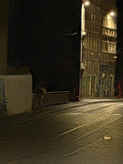 The image depicts a street scene at night in Ghent, Belgium. In the foreground, tram tracks run diagonally across a cobblestone street, illuminated by streetlights. To the left, a white, textured wall with a fence behind it is visible. A bicycle is parked near the wall. In the middle ground, two people, a man and a woman, are walking away from the viewer. The man appears to be wearing a yellow top and dark trousers, while the woman is wearing a dark top and lighter trousers. They are walking alongside a railing. Behind them, a multi-story building with numerous windows and arched detailing is illuminated. The building's facade appears to be made of stone or brick. The sky is dark, indicating nighttime.