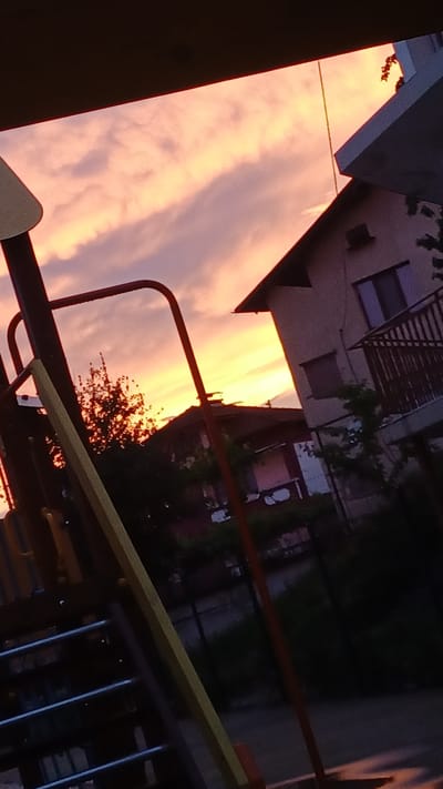 This image depicts a view of the sky during sunset in the town of Ablanitsa, Bulgaria. The sky is filled with dramatic clouds colored in hues of orange, yellow, and pink, suggesting the sun is low on the horizon. In the foreground, a playground structure, characterized by its red and yellow metal bars, is partially visible. To the right of the playground, two houses are silhouetted against the colorful sky. The house closer to the foreground has a distinct angled roof and a balcony with railings. The overall scene is one of a suburban environment transitioning into evening.