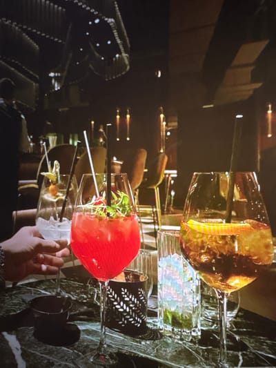 The image shows a close-up of a hand holding a cocktail with a red garnish, possibly a strawberry. The cocktail is served in a tall glass with a thin stem, resting on a multi-tiered glass stand. Another cocktail, appearing amber in color, is visible in a similar glass above and to the left of the main subject. The background is dark and out of focus, with reflections on a marble-like surface and hints of what might be a bar or restaurant setting. The lighting suggests an indoor environment, possibly at night or in a dimly lit space. The focus is on the drinks and the hand, creating an intimate and possibly sophisticated atmosphere. No visible text is present in the image. The location context of Pori, Finland, does not provide specific visual cues within the image itself.