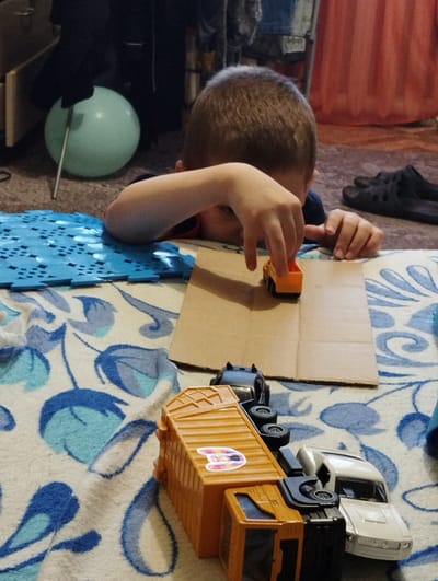 A young boy is playing with toy cars on a carpeted floor. He is sitting down, with his upper body partially obscured by his arm as he focuses on a small orange toy truck. In front of him is a piece of cardboard, serving as a road for the toy vehicles. Behind the orange truck, other toy vehicles are lined up, including a white car and a larger orange and black garbage truck. To the left of the boy, a light blue balloon is propped up against a piece of furniture. The setting appears to be indoors, likely a living room or play area, with a patterned rug and a glimpse of curtains in the background. The lighting suggests it might be daytime. There is no visible text in the image.