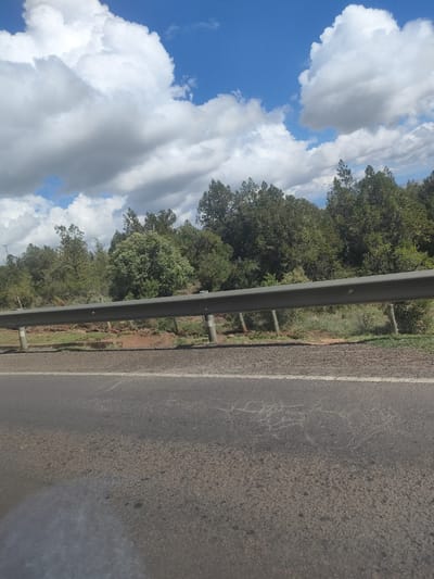 The image depicts a roadside scene in the Kisima ward of Kenya. A two-lane asphalt road with a white line divider is in the foreground. A grey metal guardrail runs parallel to the road, separating it from an area of red soil and green vegetation. Beyond the vegetation, a line of green trees is visible. The sky is blue with scattered white clouds. No people are visible.