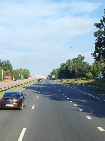 The image depicts a highway scene with a clear blue sky and scattered clouds. The road is asphalt with white dashed lines marking the lanes. Several vehicles are visible: a brown sedan in the foreground, a dark sedan further ahead, and several trucks, including a red one and an orange one, on the left side of the road, possibly on an adjacent lane or shoulder. The right side of the highway is bordered by trees and vegetation, with a blue traffic sign indicating lane guidance. The overall impression is a sunny day on a highway in a rural or semi-rural area. The location is identified as Pribrezhnyi, Russia. There are no discernible people in the image.