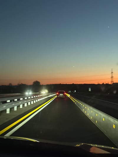 The image depicts a highway at dusk, likely in Germany. A vehicle is traveling on the road, indicated by its headlights and taillights visible in the distance. The sky transitions from a dark blue to a yellow-orange hue near the horizon. An electrical tower is visible in the background. The road is marked with yellow and white lines, and a guardrail is present along the side. Yellow reflective lights line the edge of the highway on the far side. The location in Aachen, Germany, cannot be determined from the provided image and requires additional information.