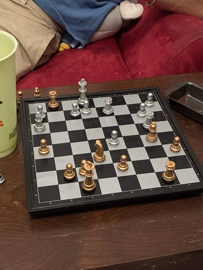 The image shows a partially visible individual sitting on a red couch next to a chess board. The chess board is set on a dark wooden surface. The chess pieces are arranged mid-game. To the bottom left of the chess board is a green container. No information suggests this image was taken in Coatzacoalcos, Mexico.
