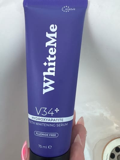 This is a close-up, eye-level, vertical shot of a tube of "WhiteMe V34 Hydroxyapatite Teeth Whitening Serum." The tube is a vibrant purple and lies horizontally across the frame. The text on the tube is white and oriented vertically, so it is readable as the tube rotates. The text includes "WhiteMe," "V34," "HYDROXYAPATITE," "TEETH WHITENING SERUM," "FLUORIDE FREE," and "75 ml e." On the left end of the tube, there's a white EU symbol with stars and the text "MADE IN EUROPE."
A person's hands with long, light-pink acrylic nails are visible on the right side of the image, slightly obscuring the end of the tube. The background appears to be a white, textured surface, possibly a sink or countertop, with a drain visible in the upper right corner, suggesting a bathroom or kitchen setting. The lighting is bright and even, with no distinct shadows, indicating an indoor setting. There are no specific time of day or weather indicators. The overall impression is a product shot with a personal touch due to the hands in the frame.