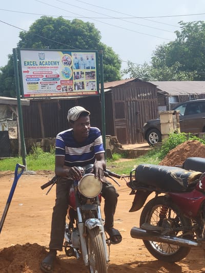 A man wearing a striped blue and white shirt, dark pants, and sandals is seated on a motorcycle next to a sign for Excel Academy in Kwamba, Nigeria. Another motorcycle is partially visible in the background. The school sign advertises primary, secondary, and pre-nursery classes, also including contact information. A corrugated metal building with a written number on it is also in the background. The ground is dirt. A blue shovel leans on the side.