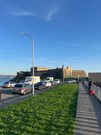 The image depicts an outdoor coastal scene in La Coruña, Spain, centered on the historic Castillo de San Antón fortification. The sky is clear blue with minimal cirrus clouds.
In the foreground, a paved stone promenade occupies the right side of the frame, alongside a strip of vibrant green grass. To the left of the grass is a paved road where approximately fifteen vehicles are parked in a single line, extending towards the castle. These include various sedans and hatchbacks, notably a grey Mercedes-Benz B-Class wagon (license plate 9507 JTF) in the foreground and two white camper vans.
The Castillo de San Antón, a large stone fortification with battlements, stands prominently in the background on a rocky promontory. A small lighthouse structure is visible on the left side of the castle. A section of a stone wall extends from the castle towards the right. Two grey streetlights with curved arms and modern fixtures are positioned along the scene.
On the paved promenade in the right foreground, a single man wearing a dark jacket stands facing the water, positioned next to a grey metal railing. A fishing rod is visible near him, resting against the railing, and an orange bag or backpack is on the ground at his feet. Several indistinct human figures are visible near the entrance area of the castle in the background. The scene is brightly lit by the sun, casting long shadows towards the right, indicating either early morning or late afternoon.