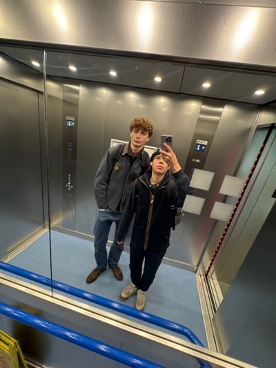 The image depicts two males in a mirror selfie taken inside an elevator, located in Newcastle upon Tyne, United Kingdom. The elevator interior features brushed stainless steel walls and a light blue floor. The male on the left, with curly brown hair and white earbuds, wears a grey work-style jacket, blue jeans, and brown shoes, with a dark backpack visible. The male on the right, wearing a dark beanie, a dark zippered jacket over a dark hoodie, dark trousers, and light-colored boots, holds a silver smartphone with both hands, angled upwards to capture their reflection. He also has white earbuds and a dark patterned backpack. Visible on the elevator walls are control panels: one on the left displaying a floor indicator "S" with an upward arrow, and another on the right featuring square buttons and a vertical red LED strip. A blue handrail is present in the lower foreground, below which a partial yellow "WET FLOOR" sign is reflected.