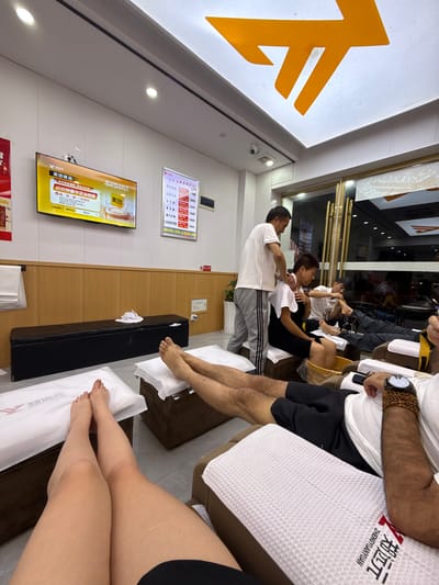 Here is a detailed description of the image:
**Scene:** The image captures the interior of a Zheng Yuang Yuan foot massage establishment in Zhangjiajie, China. The space appears to be clean and orderly, with multiple massage chairs or benches positioned for clients.
**People:** Several individuals are visible in the scene.
* Massage Therapists: There is at least one massage therapist shown attending to a client. The therapist is dressed in a white shirt and grey sweatpants.
* Clients: Several clients are visible, seated in the massage chairs/benches. One is receiving a neck massage.
* There are at least two other clients whose legs are visible.
**Objects:**
* Massage chairs/benches: The primary furniture is the massage seating, upholstered in white and brown.
* Towels: There are clean, folded white towels on the seats/benches. The Zheng Yuan Yuan logo is visible on the towels.
* Television Screen: A television on the wall displays an advertisement for a 65-minute deluxe foot massage. The ad features branding for "Meituan" (a Chinese e-commerce platform).
* Menu Boards: Small menu boards hang on the wall, presumably listing the various massage services and their prices.
* Other Items: There are black side tables near the massage chairs and various items such as the brand logos and a security camera.
**Actions and Interactions:**
* Massage Therapy: The key action is the massage therapy being administered. One client is having their neck massaged.
* Relaxation: Clients appear to be in a relaxed state, receiving foot or neck massages.
**Environment:**
* Lighting: The space seems well-lit with overhead fluorescent lights.
* Cleanliness: The interior appears clean and well-maintained.
* Atmosphere: The atmosphere suggests a comfortable and relaxing environment designed for massage and foot care.