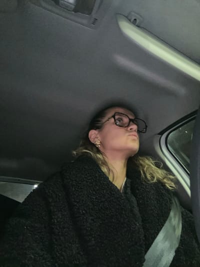 The image captures the interior of a vehicle during low light conditions. A female individual is seated on the right side of the frame, facing partially away from the camera and looking upwards and towards the left. She is wearing dark-framed eyeglasses and a gold-toned, intricate earring on her right ear. Her hair is light-colored and wavy, with some strands visible around her shoulders. She is dressed in a dark, textured coat or jacket over a white garment, and a light-colored seatbelt is fastened across her chest from her right shoulder. The vehicle's light grey headliner, an overhead console, and a light-colored interior pillar or grab handle are visible. The external environment through the window on the far right is dark and indistinct. No specific landmarks or features are present within the image to identify a precise location within the city of Saint-Étienne, France.