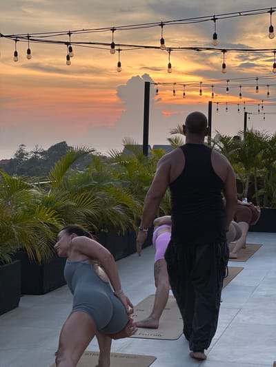 A medium shot depicts a yoga session in Tibubeneng, Indonesia, possibly on a rooftop, given the elevated perspective and view of the sunset. Four individuals are visible; one male is wearing a black tank top and black pants, positioned in the center, engaging in a yoga pose. A woman in grey workout clothes is also practicing yoga. Another figure is on a mat in the background, wearing pink pants. Strings of outdoor lights hang above, against a backdrop of a sunset sky with hues of orange, yellow, and grey clouds. Palm trees are interspersed throughout the scene. The floor is a light grey material, likely concrete or tile.