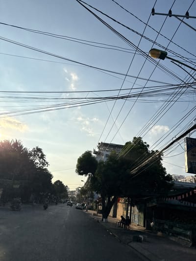 The image depicts a street scene in Bac Nha Trang Ward, Vietnam, likely during the late afternoon or early evening, as indicated by the low sun angle and long shadows. The sky is a pale blue with scattered white clouds. A prominent feature of the image is the dense network of overhead power lines crisscrossing the frame, intersecting with a streetlamp.
The street itself is paved and appears to be a two-way road, with some vehicles, including motorcycles and cars, visible in the distance. The left side of the street is lined with trees and some low-rise structures, while the right side features buildings, some with storefronts. Two individuals are seated on chairs outside a building on the right side of the street, casting long shadows. The overall impression is of a typical urban street in Vietnam, characterized by its dense infrastructure and daily life.