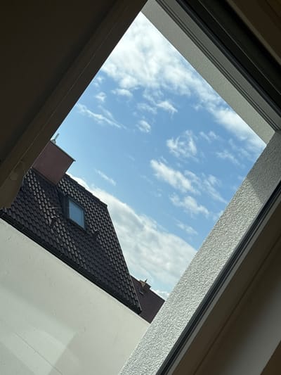 The image presents an exterior view observed through an angled window frame, likely a skylight, from an indoor position within Germering, Germany. The upper and right sections of the frame display a blue sky dotted with scattered white cumulus clouds. In the lower-left, a segment of a residential building is visible, featuring a steeply pitched roof clad in dark brown, overlapping tiles. A small, rectangular window is integrated into this roof. A reddish-brown chimney section is discernible above and slightly behind this roofline. Beneath the dark tiled roof, a substantial, unadorned white wall of a structure extends across the lower-left. A portion of another dark tiled roof with a small, indistinct architectural feature on its peak is visible in the distant background. The scene is illuminated by ambient daylight, indicating fair weather conditions. No individuals, actions, or interactions are present.