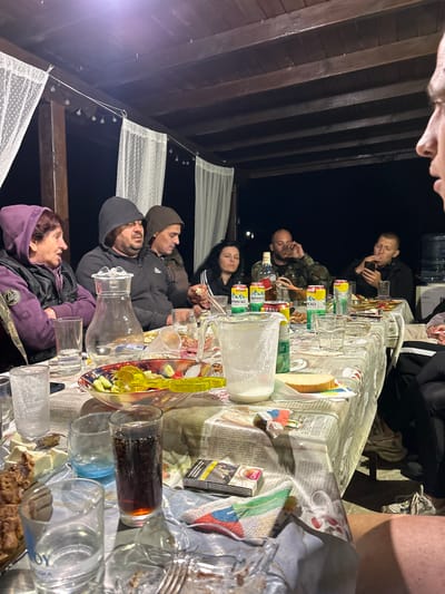 The image depicts an outdoor gathering around a table, possibly on a porch or patio, with a wooden overhead structure and draped white curtains. The setting appears to be evening or night, with artificial lighting present. Several people are seated around the table, engaged in conversation or eating.
On the table, there are various objects, including a glass pitcher, individual drinking glasses (some containing liquids), a bowl of salad, a plate with food items, cans of soda, a bottle of alcohol, and a pack of cigarettes. The table is covered with a patterned tablecloth.
In the foreground, a person's legs are visible, suggesting they are seated at the table and the photo is taken from their perspective. The people in the background are partially obscured by the table and other individuals.
The image does not contain any specific elements that would directly indicate a location in the city of Gabar, Bulgaria. The scene is generic and could represent a similar gathering in many locations.