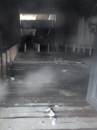The image depicts the interior of a dark, possibly abandoned structure. The floor is made of wooden planks, some of which appear worn and dirty. Several wooden posts are visible, arranged in rows extending into the depth of the space. The environment appears to be an industrial or storage area, with a bright opening at the far end suggesting an exit or a light source. Debris is scattered across the floor, including small white objects and what looks like fragments of plaster or concrete. There is a general atmosphere of disrepair and neglect. The location is identified as Shipka, Bulgaria.