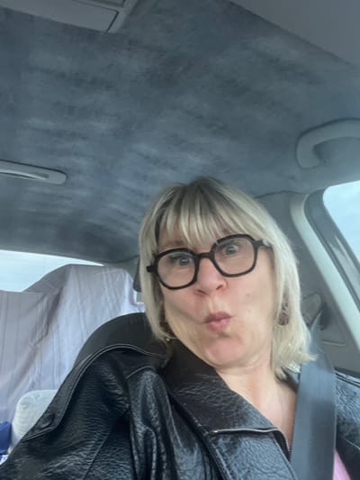 The image displays a female individual situated within the passenger compartment of a vehicle. The subject has blonde hair with bangs, wears dark-rimmed hexagonal glasses, and exhibits a 'kissing' or puckered facial expression with wide-open eyes. She is dressed in a black, textured jacket, with a portion of a pink garment visible beneath, and is secured by a dark seatbelt across her right shoulder and chest. A dark, possibly reddish, earring is visible on her right earlobe. The vehicle's interior includes a grey, textured headliner, an overhead console towards the top center, and a grey grab handle on the upper right side. On the left, a light-colored, striped fabric covers part of the rear seating area or cargo space, visible through a window that reveals a blurred, bright outdoor environment. The specific external geographic location in the city of Sauzet, France, cannot be determined from the visual content of the image.