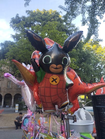In Lugano, Switzerland, on a bright day with a clear sky, a large inflatable figure resembling the cartoon character Bing Bunny is prominently displayed outdoors, likely at a street market or fair. The balloon is black and orange with a plaid-patterned red outfit, and it is attached to a stand with several other balloons, some of which are metallic silver and pink. Behind the balloons, lush green trees form a dense canopy, and in the background, a building with arched windows and a fountain can be seen. To the left of the display, a man is crouched down, smoking what appears to be a cigarette. The overall scene suggests a lively, possibly festive, outdoor environment.