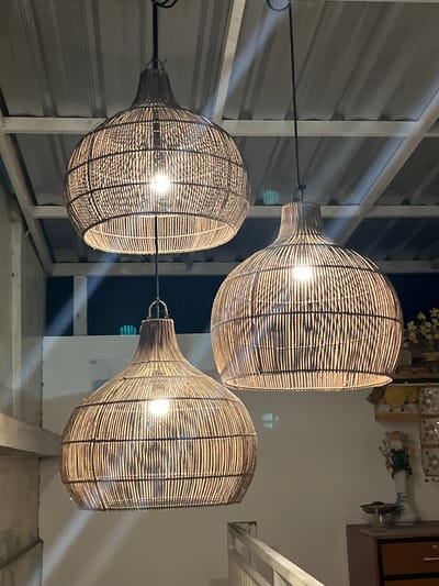 The image shows three spherical lamps hanging from the ceiling, likely in a building in or near Desa Delod Peken, Indonesia. The lamps are made of woven material. The image is taken from below, looking up at the lamps. The background includes a corrugated metal ceiling, structural beams, and a glimpse of a room with furniture and decorations. The lights within the lamps are illuminated.