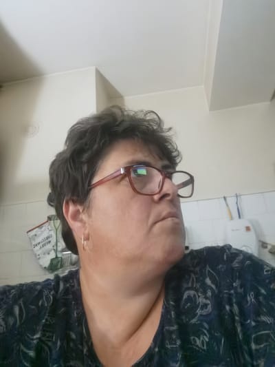 This is a close-up, medium shot of a middle-aged woman with short, dark, curly hair, wearing red-rimmed glasses and a dark patterned shirt. She is looking to her right, away from the camera. The background appears to be a tiled kitchen or bathroom wall, with a white object and two toothbrushes visible on a shelf. A bag with the words "MERRY CHRISTMAS" printed on it is hanging on the wall to the left of the woman. The lighting is bright, and the scene is likely indoors. The location is not specified as being in Lomets, Bulgaria in the image.