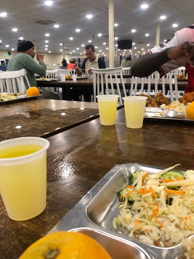 The image depicts an indoor cafeteria or large dining hall located in النعيلة, Saudi Arabia. The scene is illuminated by numerous overhead recessed circular lights set into a white ceiling, supported by light-colored pillars.
In the foreground, a glossy, dark brown table surface exhibits visible liquid spills and reflections. A portion of an orange is present at the bottom left. A clear plastic cup filled with a light yellow liquid is situated on the table, along with a section of a metal cafeteria tray containing a mixed salad composed of shredded cabbage, carrots, and sliced cucumbers.
In the midground, another section of the same glossy table holds two additional clear plastic cups containing light yellow liquid. To the right, a metal cafeteria tray displays several pieces of fried chicken, a portion of salad, and an orange fruit. A person with dark skin, wearing a red short-sleeved shirt with white and navy blue accents, has their arm and hand extended towards this tray.
The background reveals multiple rows of dark tables paired with white-backed wooden chairs. Several individuals are seated at these tables, engaged in eating. A man on the left, wearing a dark beanie and a light green shirt, is actively eating from a plate with a fork, with an orange visible on his table. In the center, another man with short dark hair, dressed in a white and navy blue polo shirt, is eating with chopsticks from a white bowl. Other blurred figures are discernible further in the background, also dining. Stainless steel equipment or cabinetry is visible along the far wall.