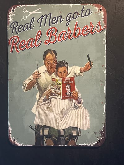 The image depicts a rectangular tin sign with a weathered, vintage appearance. The sign features a faded light blue-gray background. On the left side, in large red and navy cursive lettering outlined in white, it reads "Real Men go to Real Barbers". A pair of scissors is illustrated next to the text.
The right side of the sign shows a vintage illustration of a barber and his client. The barber is shown holding a straight razor. The client sits in a barber chair covered in a striped barber cape and is reading a comic book. He is also holding a book titled "Roger Wilkerson". The illustration has a slightly distressed, aged aesthetic.
The sign has a rusted-look trim with small holes in each corner, presumably for mounting. The environment appears to be a dark, neutral surface upon which the sign rests.
There is no information to suggest that this image is located in the city of Selyatino, Russia.