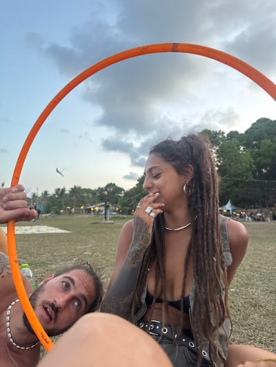 This image depicts two individuals in Ban Pang Pring, Thailand, set against a grassy outdoor area at what appears to be late dusk or early evening. The sky is overcast with a mix of blue and grey clouds, and a distant, dark, kite-like object is visible.
In the foreground, an orange hoop frames the upper body of a female and the head of a male. The female, positioned on the right, has dark skin, dreadlocks pulled back into a high ponytail, a nose ring, large silver hoop earrings, a silver choker, and multiple silver rings on her left hand. Her left forearm is heavily tattooed with a mandala-like pattern and solid black ink extending upwards. She wears a black bikini top and a grey, possibly denim, sleeveless vest. Her expression is one of amusement or laughter, with her eyes closed or looking down, and her right hand partially covering her mouth. Below her, a black item of clothing, possibly shorts or a skirt, with metal eyelets or studs, is visible.
To the lower left, a male with light skin, dark hair pulled back, and a beard is visible, lying on his back. He wears a necklace made of white shells. His left arm is holding the orange hoop. His head is tilted upwards, mouth slightly agape, and he appears to be looking towards the female or the sky. A green-capped plastic water bottle rests on the grass near his arm.
In the background, a line of trees is visible. Beyond the trees and across the grassy field, indistinct structures, possibly huts or tents, are illuminated with artificial lights, suggesting a populated area or ongoing event.