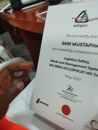 The image shows a certificate presented to "Sani Mustapha" for completing a "Logistics Safety Minds and Management System" training program, certified by "Safety Plus", with "ISO 39001:2012/SPSCAT-NG Top" standards. The certificate was issued in May 2025 by Lafarge. The certificate is held in a person's hand. The coordinates given (latitude: 10.929, longitude: 11.476) are in Ashaka, Nigeria.