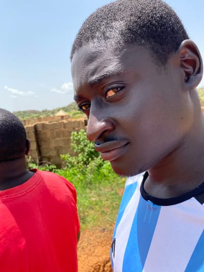 The image is a close-up, outdoor portrait of two young Black males in Barakin Angundi, Nigeria. The primary subject, in the foreground and facing the camera, is a male with dark skin and short, dark, curly hair. He is wearing a blue and white striped jersey with a black neckline. His head is tilted slightly, and he is looking towards the camera with a subtle smile. His right eye is visible, and the light reflects off his forehead and cheek. Behind him and to the left, a second male is seen from the back, wearing a bright red t-shirt. The background consists of a low, reddish-brown brick wall, lush green foliage, and a hazy sky with some clouds. The ground appears to be a mix of reddish-brown soil and dry grass. The overall impression is a casual, sunny outdoor setting.