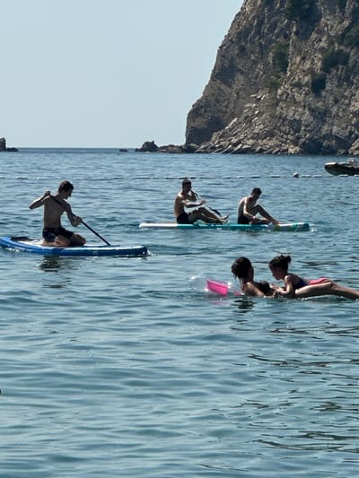 The image shows a sunny day at the beach in Đurovići, Montenegro. In the foreground, two young girls are playing in the water on inflatable toys. Behind them, three young men are paddleboarding. The man on the left is kneeling and paddling, while the other two are sitting on their boards. In the background, a large rocky cliff rises from the sea. The water is a clear blue, and the sky is a pale blue. The scene is peaceful and idyllic, capturing a typical summer day at the beach.