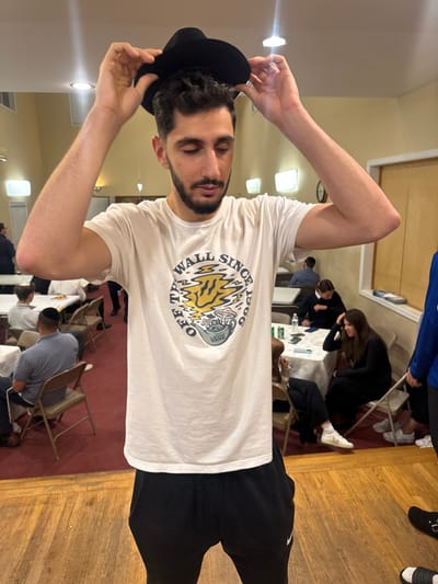 This image depicts an indoor gathering in Bergenfield, United States. In the foreground, a light-skinned male with dark, curly hair and a beard stands centrally, looking downwards. He wears a white short-sleeved t-shirt with a "Vans Off The Wall Since 1966" graphic featuring a melting smiley face and mushrooms, black sweatpants with a visible Nike logo on the left leg, and partially visible white sneakers. He is holding a black fedora-style hat above his head with both hands.
The environment features a wooden floor in the immediate foreground, transitioning to a red carpeted area in the mid-ground and background. The walls are light beige. Overhead fluorescent lighting fixtures and some recessed lights illuminate the room.
In the background, numerous individuals are seated at rectangular tables covered with white cloths, using brown folding chairs. On the left side, several men are seated, some wearing yarmulkes (kippot). On the right side, multiple people are seated, including a woman with long dark hair wearing a black long-sleeved dress, resting her head on her hand. Beverages in cans and bottles, as well as plates, are visible on the tables. A circular wall clock with a white face and black numerals is present on the right-side wall.