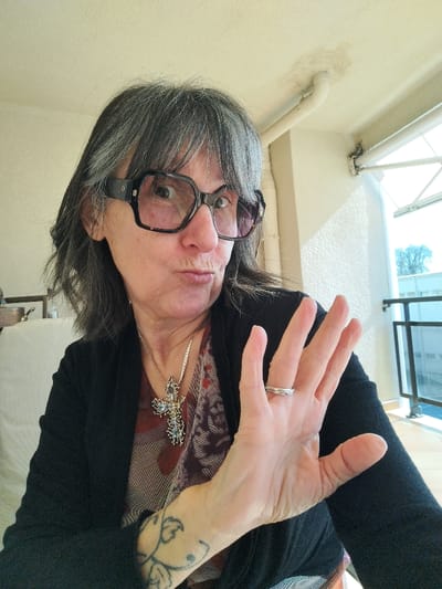 An adult female is depicted indoors in Montélimar, France, presenting a direct gaze towards the camera. She has gray hair with bangs and wears large, dark-framed eyeglasses. Her lips are pursed, and her left hand is raised with the palm facing forward and fingers splayed. A silver-colored ring is visible on her left ring finger, and a faded tattoo is present on her left inner forearm. She is dressed in a dark cardigan over a patterned top and wears a necklace with a large, ornate cross pendant featuring multiple light-colored gemstones. The indoor setting features light-colored walls and a ceiling with a visible water stain and exposed piping. A balcony with dark railings is visible through a window, offering a view of bare trees and distant light-colored structures under natural light.