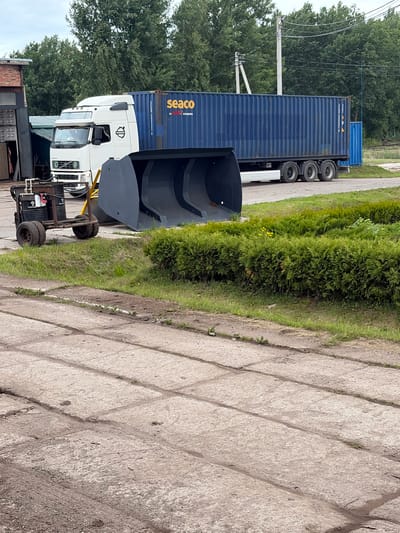 The image shows a white Volvo truck pulling a blue shipping container with "seaco" branding. In front of the truck is a large, grey industrial bucket. To the left of the truck is a small, industrial cart with two barrels. The scene appears to be in an outdoor industrial or storage area. The ground is paved with concrete slabs, and there is grass and trees in the background. The location is Zarech'e, Russia.