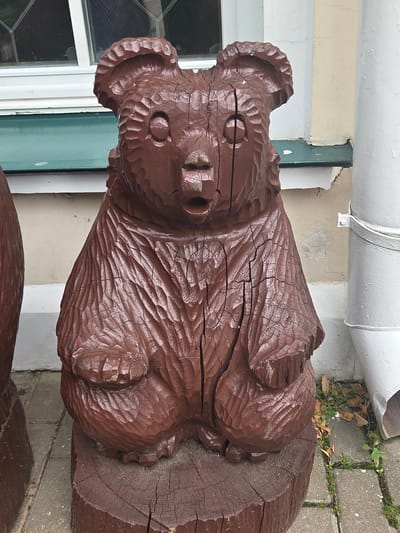 The image shows a close-up, low-angle view of a wooden sculpture of a koala, painted brown. The sculpture is detailed, with carved fur texture and distinct facial features, including round eyes and a prominent nose. It appears to be an outdoor decoration, positioned against a building with a cream-colored wall and a green trim near a window. Part of a white pipe is visible above the sculpture. The sculpture is resting on a paved area with visible grass growing between the paving stones, suggesting an exterior setting. The lighting and shadows suggest it could be daytime, but no specific time is discernible. There are no people or text visible in the image. The overall impression is of a whimsical, handcrafted garden or yard ornament. The location context provided is Sergiyev Posad, Russia.