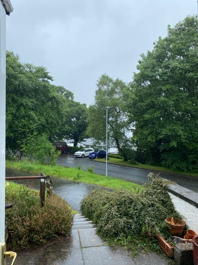 The image shows a view from a house located in Keswick, United Kingdom (latitude: 54.6, longitude: -3.12). The scene is a residential street with a few cars parked along the curb. There are lush green trees and bushes lining the street, and the weather appears overcast and wet. The ground is wet and covered in puddles. A small set of concrete steps leads down from the house to the street. There is a railing to the left of the steps. A brown pot containing a plant sits on the ground next to the bushes. The street is wet and empty. The image was captured from a window.