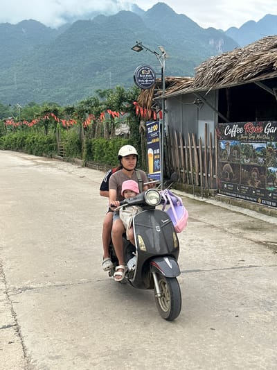 In Mai Chau, Vietnam, two individuals are riding a scooter on a paved road. The person in front, wearing a white helmet and a brown shirt, is operating the scooter. Behind them, a child wearing a pink hat is seated, also wearing a light-colored top. Both are looking forward. The scooter is dark gray with a white front fender and a silver trim. A purple and pink bag is attached to the back of the scooter. In the background, lush green mountains rise under a cloudy sky. A building with a thatched roof is visible to the right, adorned with red flags and signage, including one that reads "Coffee Rose Garden." The overall scene depicts a common mode of transportation in a rural Vietnamese setting.