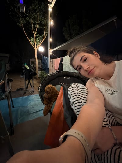 A low-angle, medium shot captures a woman and a small, brown poodle in an outdoor setting at night, likely a campground. The woman, in the foreground, is looking at the camera with her hair pulled up in a bun and is wearing a white t-shirt with the text "California Los Angeles Est. 1996". She is resting her arm on her lap, which is covered by black and white striped pants. A light-colored bracelet adorns her wrist. To her left, a poodle sits inside a dark-colored carrier. In the background, a tree with sparse leaves is illuminated by a white light at its base and a blue light fixture hanging from its branches. String lights are visible above, and laundry is hanging on a line behind the tree. Camping equipment, including chairs and what appears to be a camper van, are present. The ground is covered with a blue tarp and a grey mat. The overall scene suggests a relaxed evening at a campsite near Πόρτες, Greece.