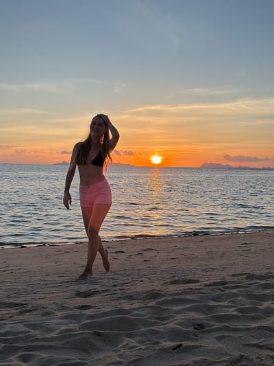 A young Caucasian woman with long, dark blonde hair is walking along a sandy beach in Baan Taling Ngam, Thailand, during sunset. She is wearing a black bikini top and pink shorts. Her left hand is raised and touching her head, and her right hand is at her side. The woman is facing the camera with a slight smile. The sun is setting over the ocean, casting a warm orange glow across the sky and water. In the distance, silhouetted islands can be seen. The sand on the beach is light brown and textured with footprints. The water is calm with small waves.
