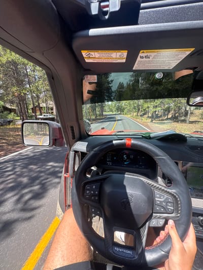 The image provides a first-person interior view from the driver's seat of a red Ford Bronco Raptor, identifiable by the "RAPTOR" emblem on the steering wheel, actively driving on a paved road in Sunriver, Oregon, United States.
**People:** One individual is operating the vehicle. Their right hand, with a short fingernail, is visible on the lower right portion of the steering wheel, holding it. Their right leg, wearing dark shorts, is visible in the lower left frame. No other individuals are discernible.
**Subjects/Objects:** The primary subject is the vehicle's interior and the road ahead. The steering wheel features black material with carbon fiber accents and red stitching, and has a distinct red center marker at the 12 o'clock position. The digital instrument cluster displays "5" (likely miles per hour) and "22" (likely degrees). The dashboard is visible, with a light-colored baseball cap resting on it. Sun visors, containing warning labels related to "HIGHER ROLLOVER RISK" and "AIR BAG," are deployed. A silver cylindrical can is wedged between the top of the windshield and the sun visor on the passenger side. The driver's side mirror reflects the road's yellow line.
**Actions/Interactions:** The individual is actively driving the Ford Bronco Raptor. The vehicle is in motion along a curved road.
**Environment:** The setting is a sunny day in a heavily forested area. The asphalt road features a double yellow center line. Tall evergreen trees densely line both sides of the road, casting shadows. To the left, off the road, a wooden cabin-style structure is visible, partially obscured by trees, with a white pickup truck parked nearby. The sky appears clear.