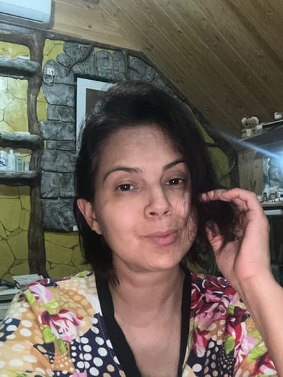 A woman is captured in a selfie, with her face filling most of the frame. She has dark hair, styled to fall around her face, and appears to be touching her hair with her right hand. Her eyes are open, and she has a slight smile on her lips. She is wearing a colorful top with a floral and polka dot pattern.
The background suggests an indoor setting with a rustic or unconventional design. To the left of the woman, a wall features a textured design resembling stone and wood, with shelves holding several bottles and containers. Above this, a white air conditioning unit is visible. The ceiling is made of wooden planks. To the right, there is a shelf with small decorative figures, possibly teddy bears, and other indistinct objects. The lighting suggests it is nighttime or the room is dimly lit, with a visible light source to the right creating a glare.
The provided information states the image's location is Kalininskaya, Russia. This detail is external to the visual content of the image itself and cannot be confirmed from the image alone.