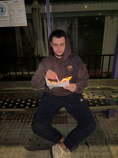 A young male is seated on a metallic, perforated bench at a bus stop in Grau I Platja, Spain, during nighttime. He is wearing a dark brown hooded sweatshirt with a Nike logo and FC Barcelona crest, dark blue Adidas track pants, and light-colored sneakers with white socks. The individual holds a white piece of paper in his left hand that is actively aflame, emitting bright yellow and orange fire. In his right hand, he holds a white cylindrical object, possibly a marker or similar implement, positioned above the burning paper. Behind him, a large glass panel of the bus shelter reflects dim interior lights and allows a view into the dark street or urban landscape beyond. To the upper left, a white sign through the glass displays "LA MARINA" and "AUTOBUS LINEA 2" with additional route information. The ground is paved with grey square tiles.