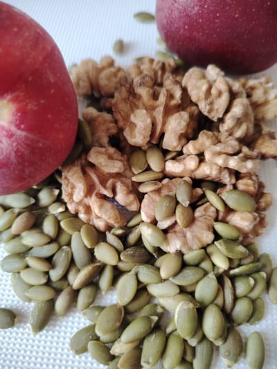 The image shows a close-up, high-angle view of pumpkin seeds, walnuts, and a red apple on a white textured surface. The pumpkin seeds are a light green and are piled in the foreground and middle ground. The walnuts are a light brown and are clustered behind the pumpkin seeds, mostly in the middle ground. A portion of a red apple is visible in the upper left corner. The lighting is bright and diffused. There are no people or animals present. The scene does not provide any information about the location in the city of Кривец, Russia.