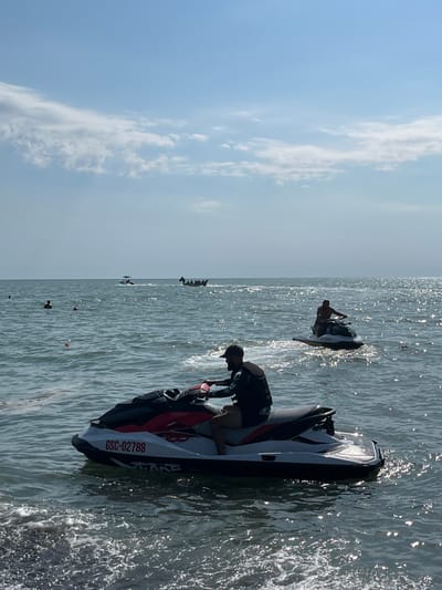 This image depicts a sunny day at the Ureki, Georgia Black Sea coast. In the foreground, a person is riding a jet ski. The jet ski is white and red with the registration number "GSC-02788" visible. Further out in the water, another person is riding a jet ski. In the middle ground, several people are visible in the water, and two boats are moving across the sea. The sky is blue with some scattered clouds, and the water is shimmering with sunlight. The overall scene conveys a recreational and leisurely atmosphere.